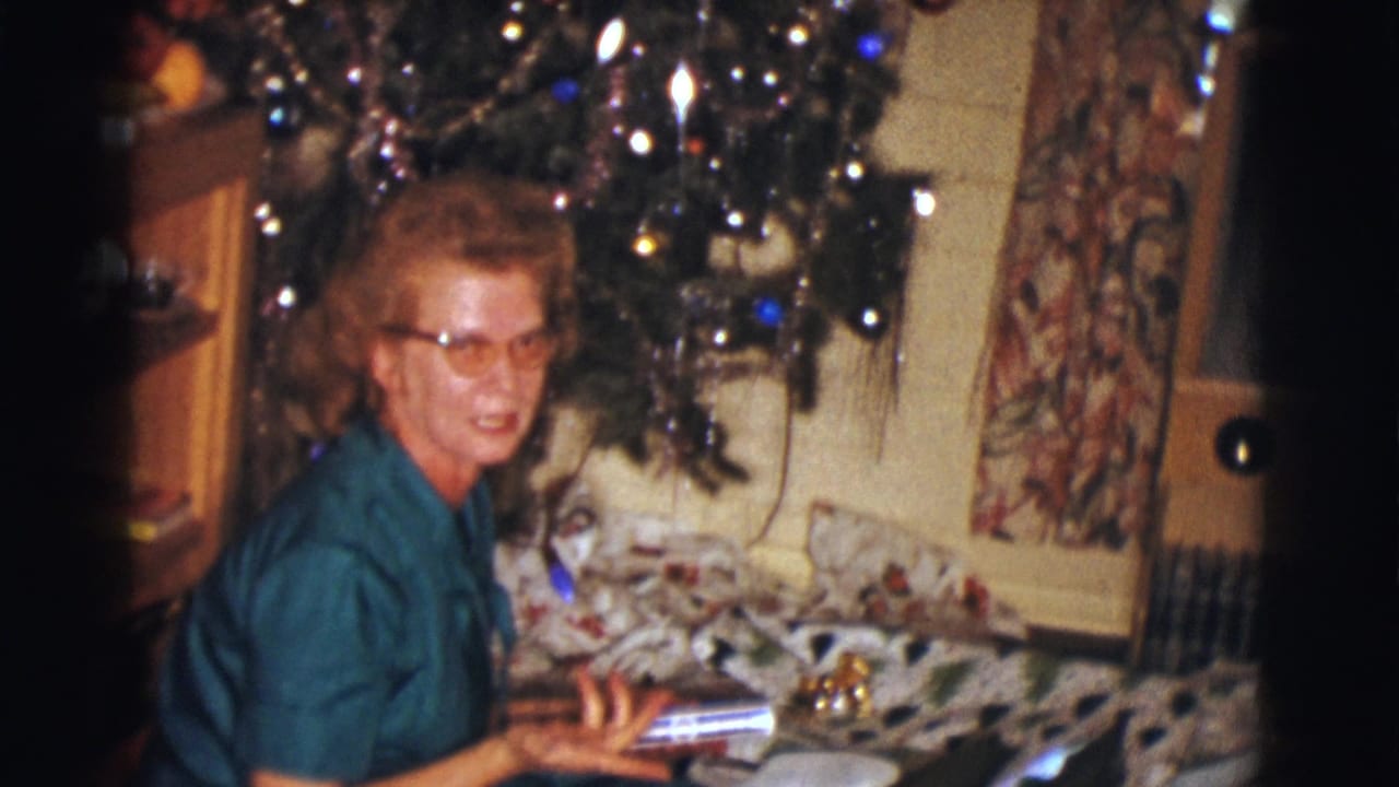 Woman in glasses sits beside a Christmas tree with wrapped gifts nearby.