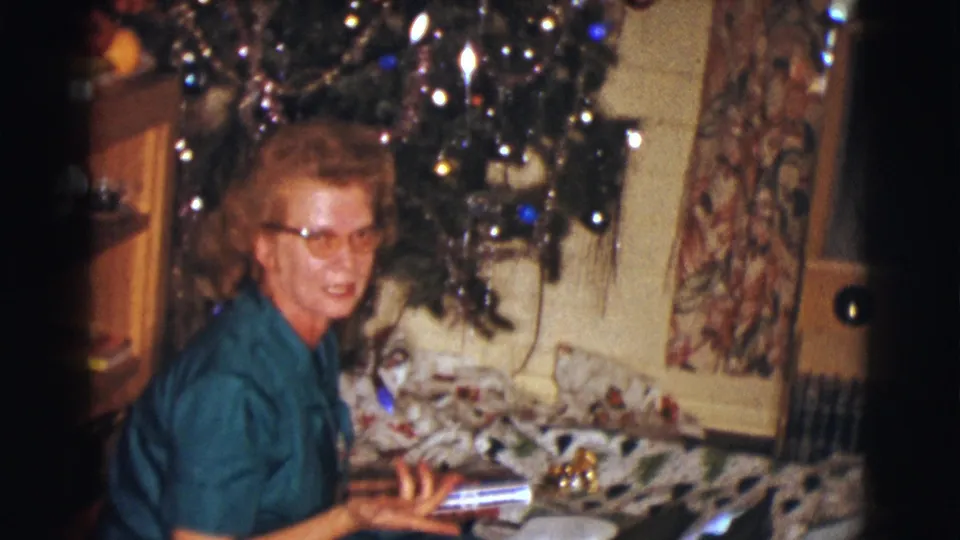 In a Clarkdale, Arizona home-movie frame from 1961, a woman in glasses sits near a decorated Christmas tree and wrapped presents.