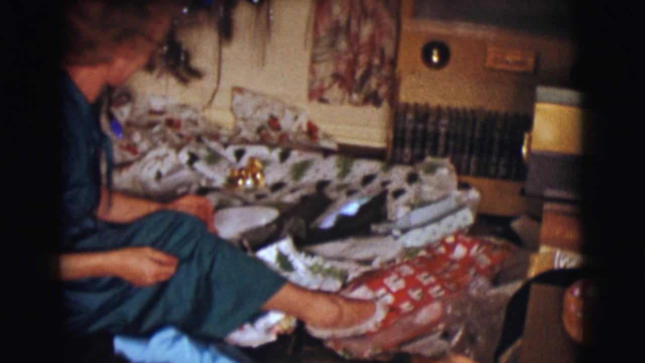 Woman in a teal dress sits beside wrapped gifts near a Christmas tree in a living room.