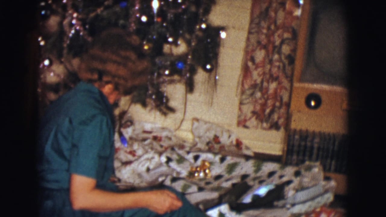 Woman sits beside a Christmas tree and wrapped presents, turned partly away from the camera.