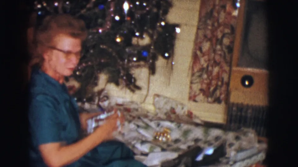 The woman, wearing glasses and a teal dress, handles a Christmas gift near the decorated tree in this 1961 Clarkdale interior.