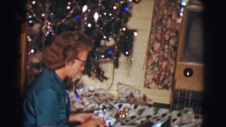 In profile, the woman sits by wrapped gifts beneath a decorated Christmas tree, with curtains and a console television visible in the home interior.