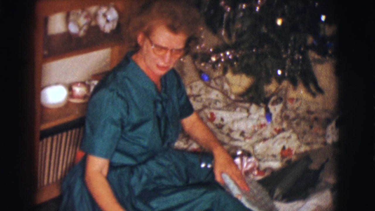 Woman in a teal dress sits beside a Christmas tree holding a wrapped present.