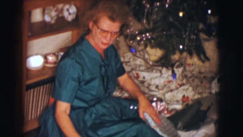 In a 1961 Clarkdale, Arizona home movie, a woman in glasses sits on the floor beside a decorated Christmas tree with a wrapped gift.