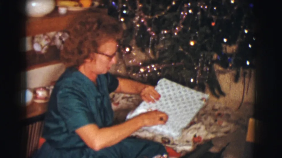The seated woman opens patterned wrapping paper on her lap near the decorated tree in the 1961 Clarkdale Christmas home movie.