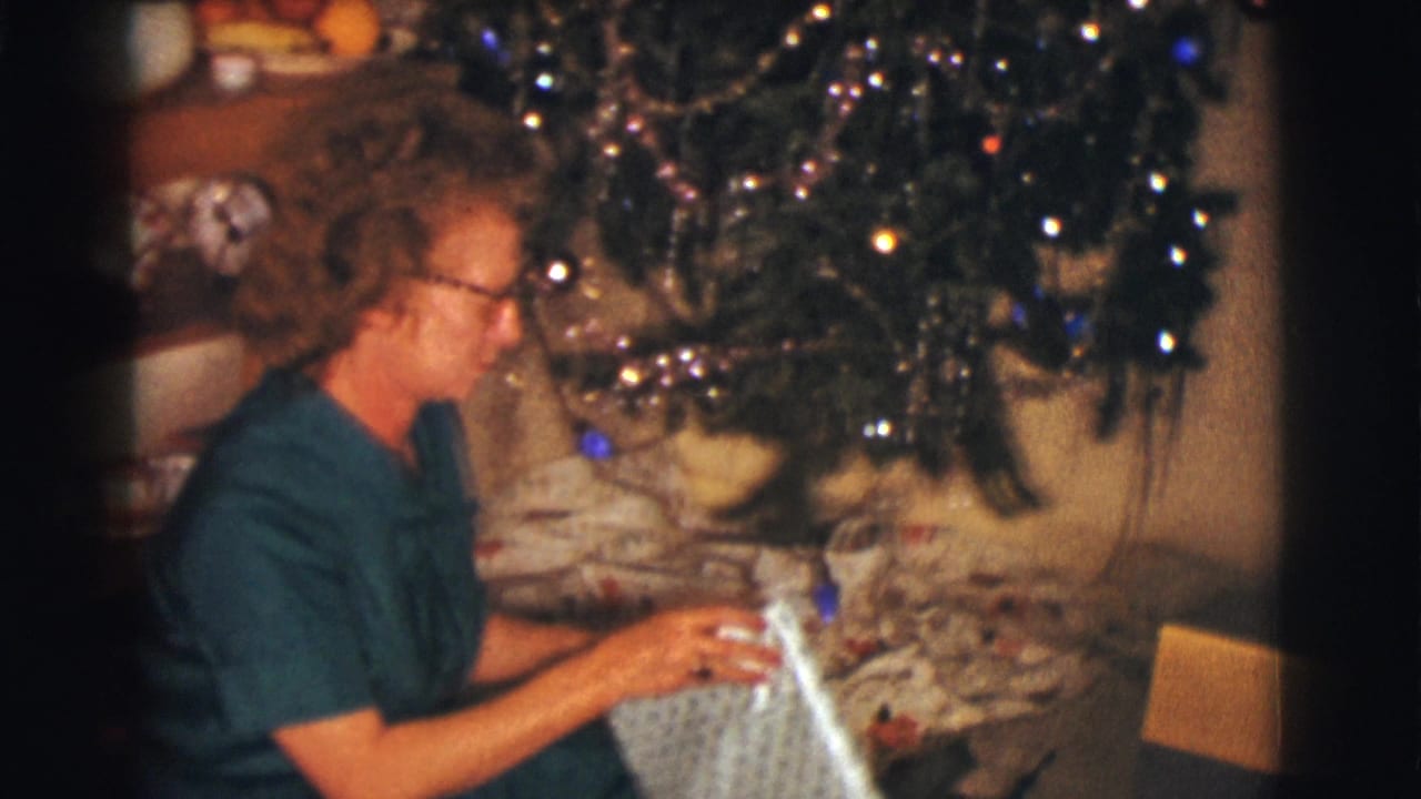 Woman in glasses sits beside a decorated Christmas tree holding wrapped paper.