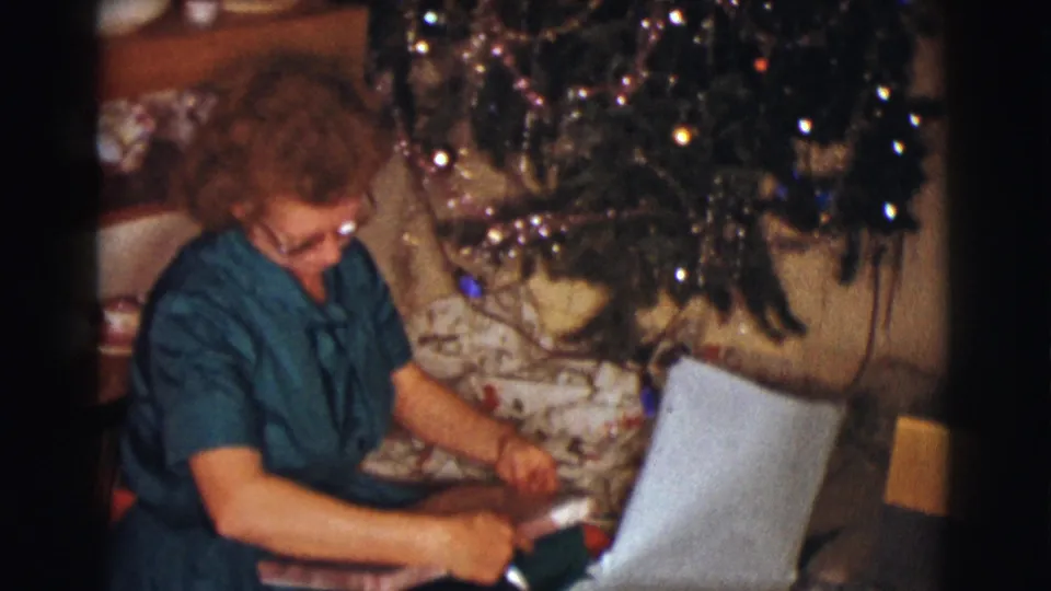 A 1961 home-movie frame shows the woman leaning over the gift area, with loose wrapping paper and the Christmas tree filling the background.