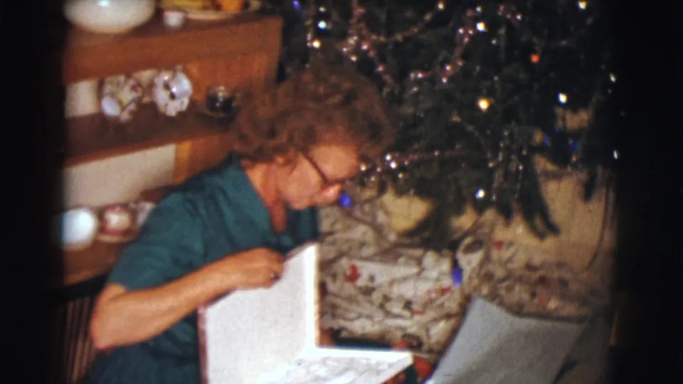 The woman raises opened wrapping paper near the Christmas tree, with ornaments and tinsel visible in the warm 8mm interior scene.