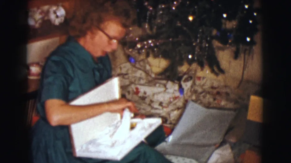 In a 1961 Clarkdale, Arizona home-movie frame, a woman in glasses sits indoors beside a Christmas tree and opens a gift box.