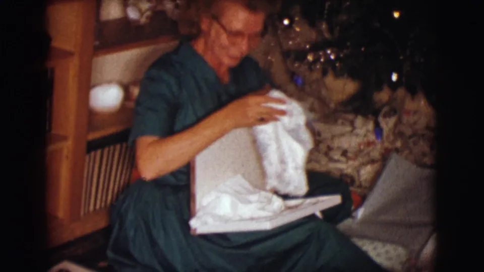 The woman in a green dress looks down at the white fabric or garment over the open box, with the decorated tree close behind her.