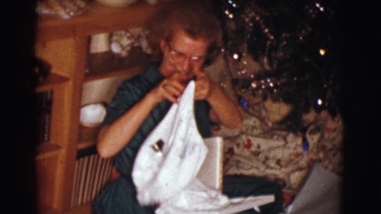 Woman in glasses opens a white Christmas gift near a decorated tree and wooden shelves.
