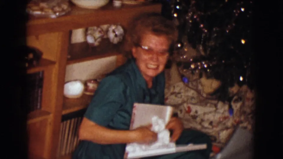 The woman laughs during the gift opening, holding a white garment or package in a decorated Clarkdale living room.