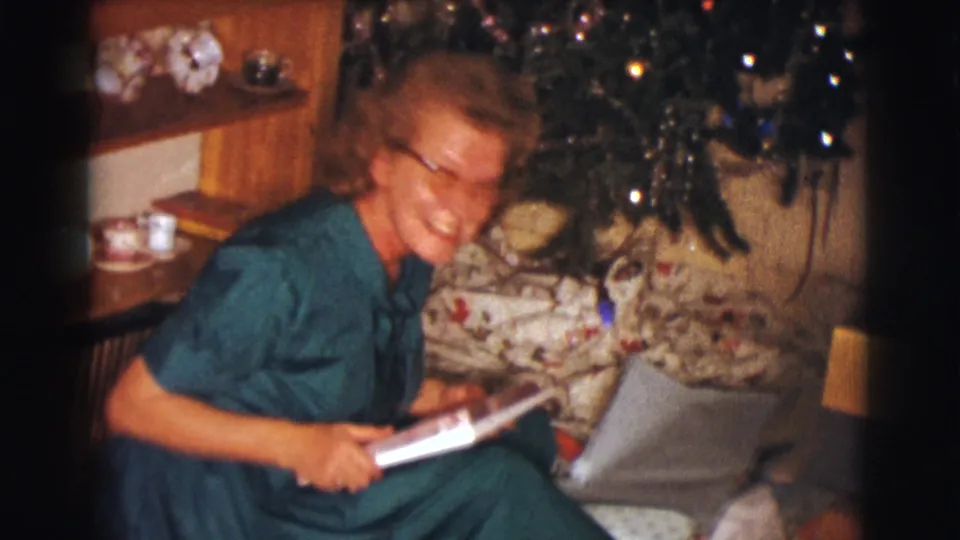A smiling woman leans forward with the opened white item in her lap during a 1961 Christmas home-movie scene.