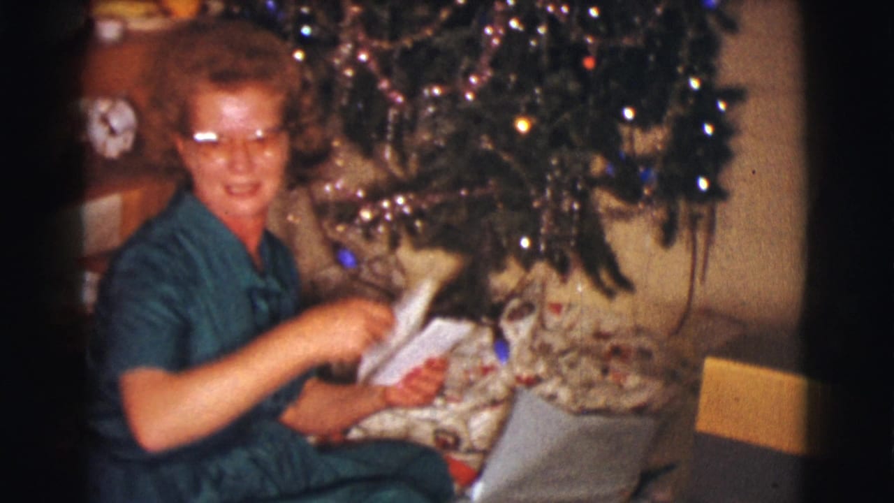 Woman seated under a Christmas tree holds white envelopes and looks toward the camera.