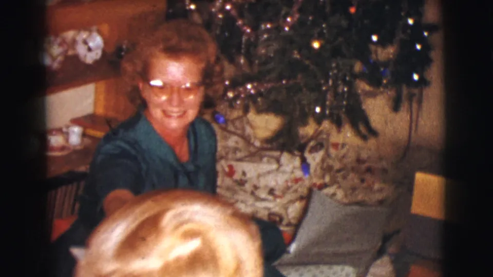 The woman smiles beside the Christmas tree while a blond child's head is partly visible in the foreground of the home-movie frame.