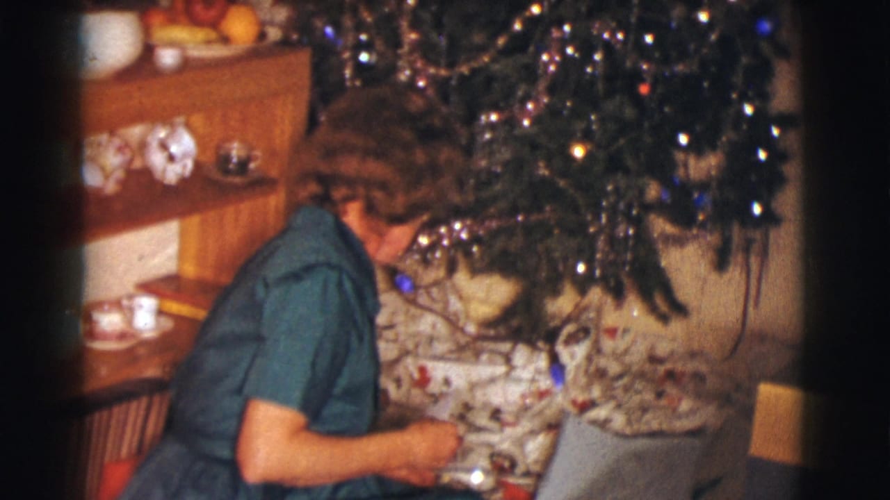 The woman leans toward Christmas presents under a decorated tree in a private Clarkdale interior, filmed on 8mm in 1961.