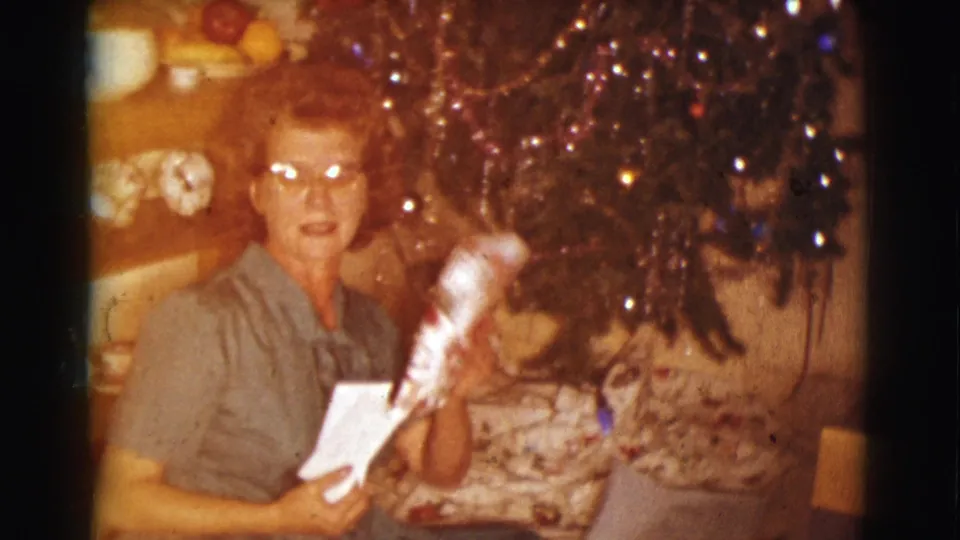 A woman wearing glasses holds a wrapped gift or holiday item near a tinsel-covered Christmas tree in 1961 Clarkdale footage.