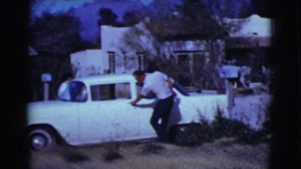 A man in a white shirt leans into a white vintage sedan outside a stucco residence in Clarkdale, Arizona home-movie footage.