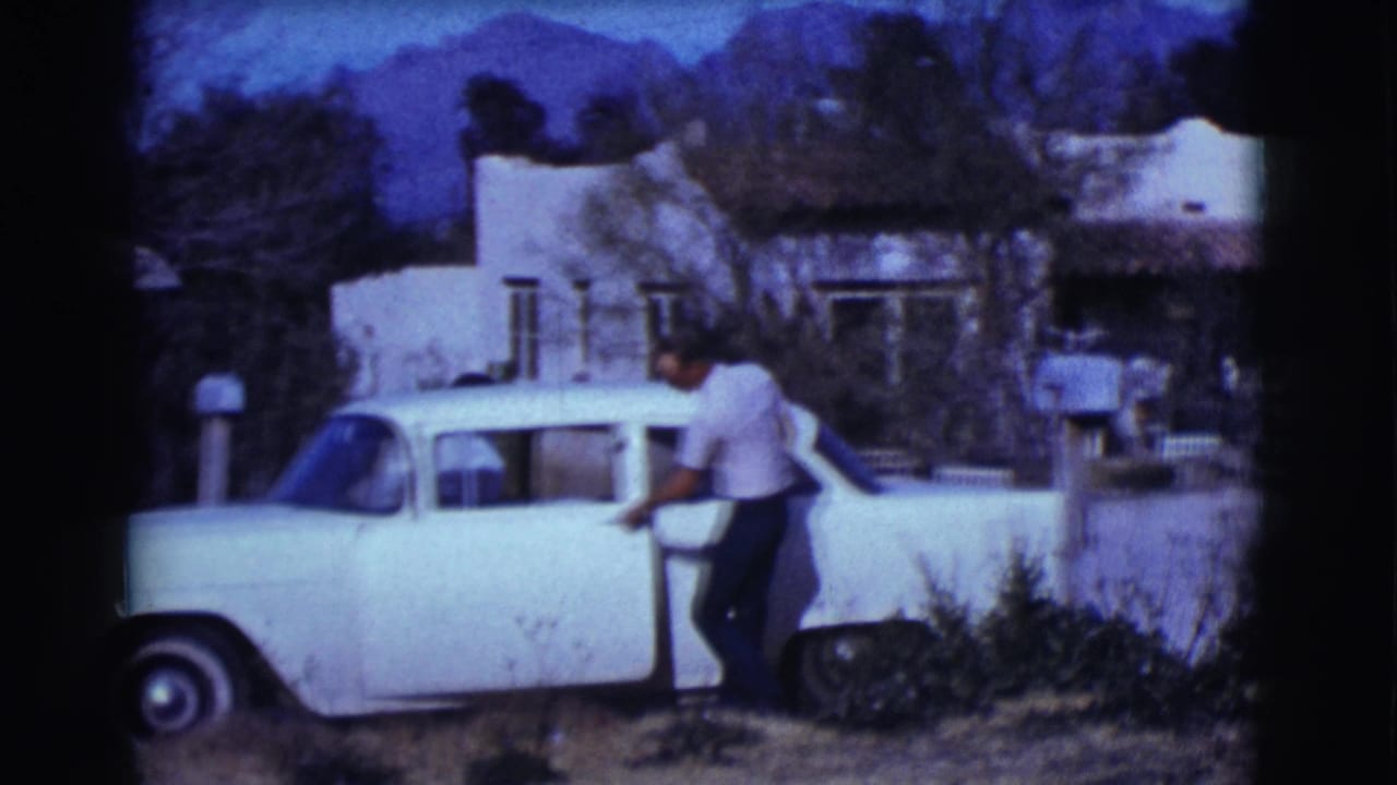 Man standing beside a white vintage sedan near a stucco house.