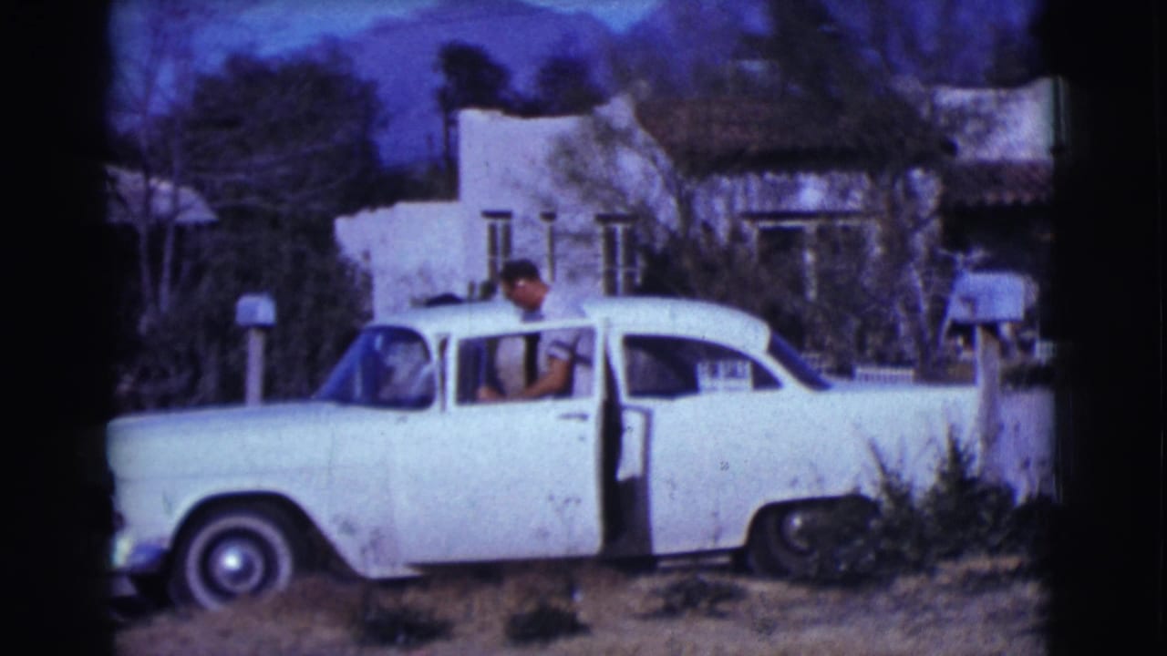 Man at the open door of a white sedan parked in front of a house.