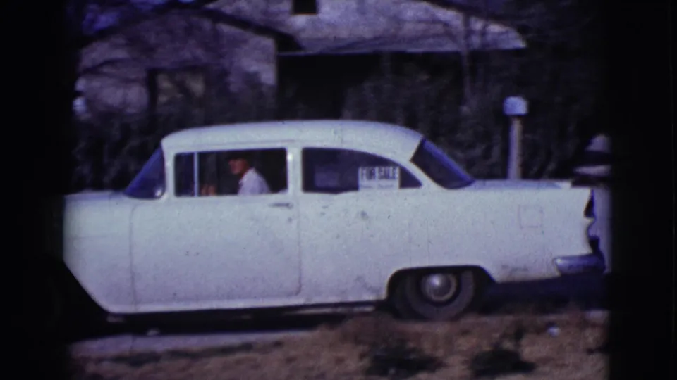 The white sedan remains parked in the residential scene, with a man visible through the side window in 1961 Clarkdale footage.