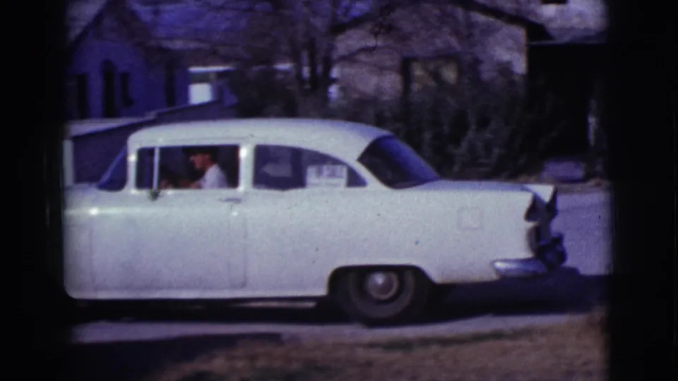 White classic sedan in a Clarkdale, Arizona residential setting in 1961, with a person visible inside and houses behind it.