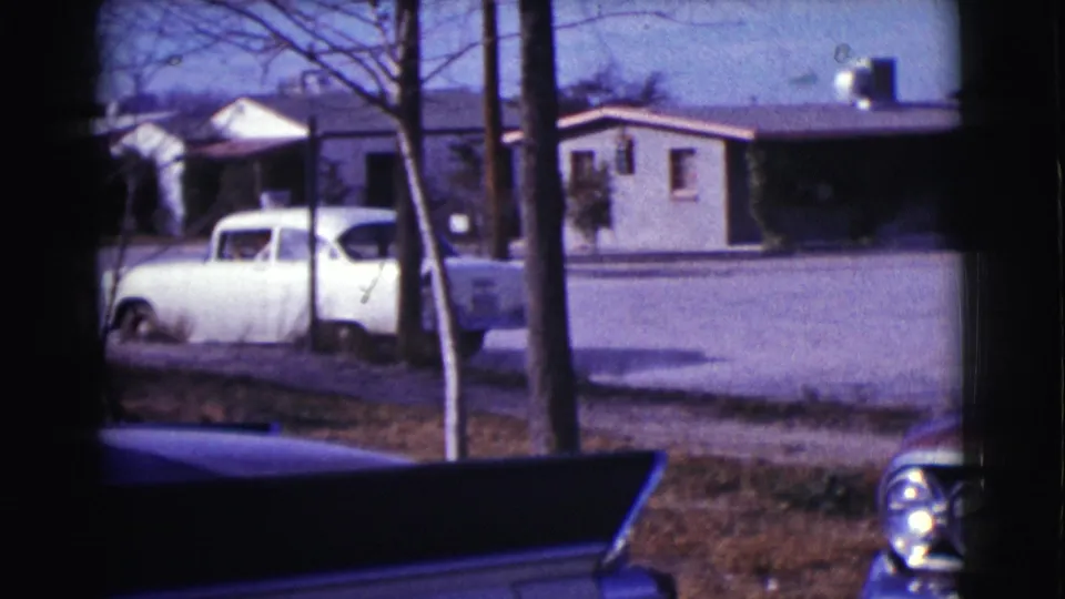 White sedan parked beyond tree trunks in the 1961 Clarkdale home-movie footage, with a house and foreground car details visible.