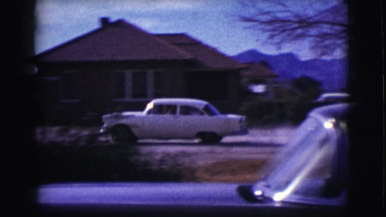 White sedan passing a residential house with distant hills behind it.