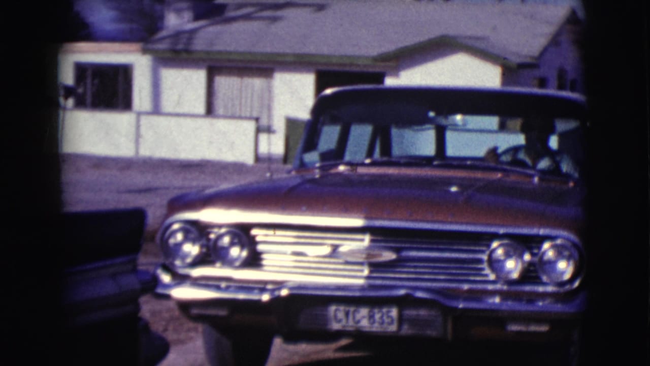 Classic sedan front with driver visible through the windshield near a house.