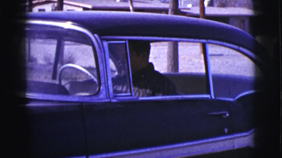 The camera frames the side of a dark mid-century sedan in Clarkdale, with a person visible through the window.