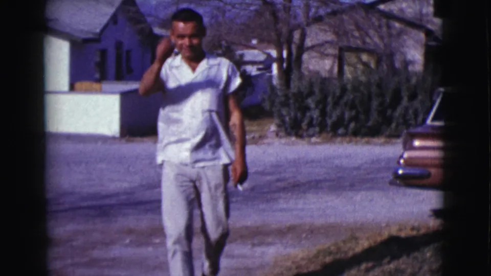 In 1961 Clarkdale, Arizona, the man walks closer to camera and raises a hand near his head, with houses and trees behind him.