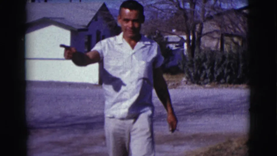 In 1961 Clarkdale, Arizona, the man points toward the camera in a sunlit residential driveway with houses and bare trees behind him.