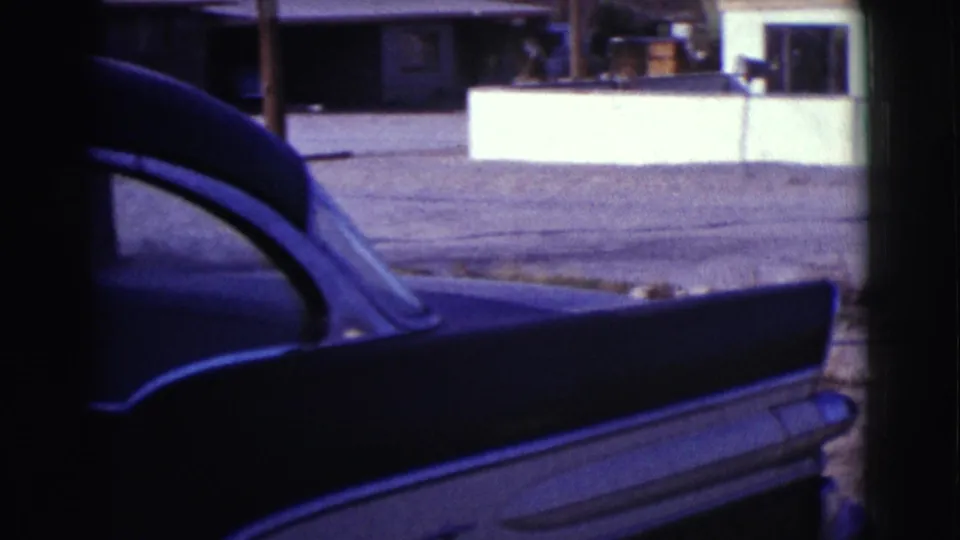 The rear side of a dark vintage car fills the foreground, with a low wall and houses behind it in the 1961 Clarkdale footage.