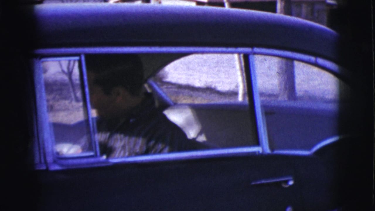 Man in a striped shirt seated inside a dark vintage car, seen through the side window.