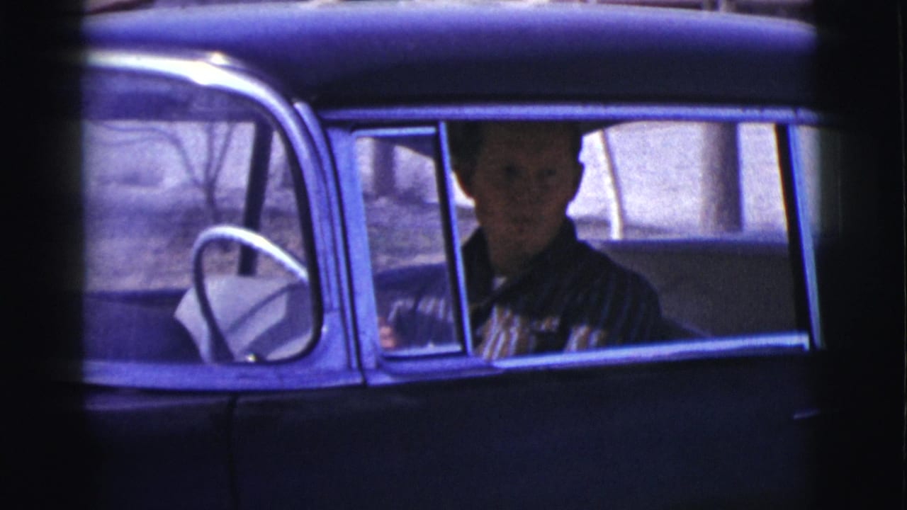 The driver looks out from the front seat of a dark vintage car in the 1961 Clarkdale home-movie footage, with the steering wheel and window frame visible.