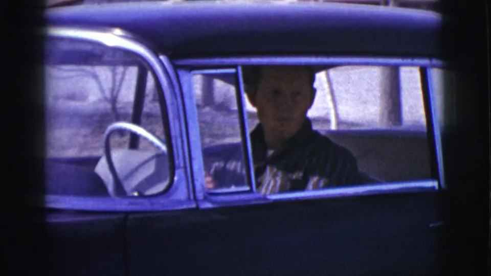 The driver looks out from the front seat of a dark vintage car in the 1961 Clarkdale home-movie footage, with the steering wheel and window frame visible.