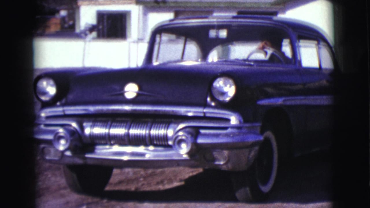 Dark mid-century car seen close from the front near a residential driveway.