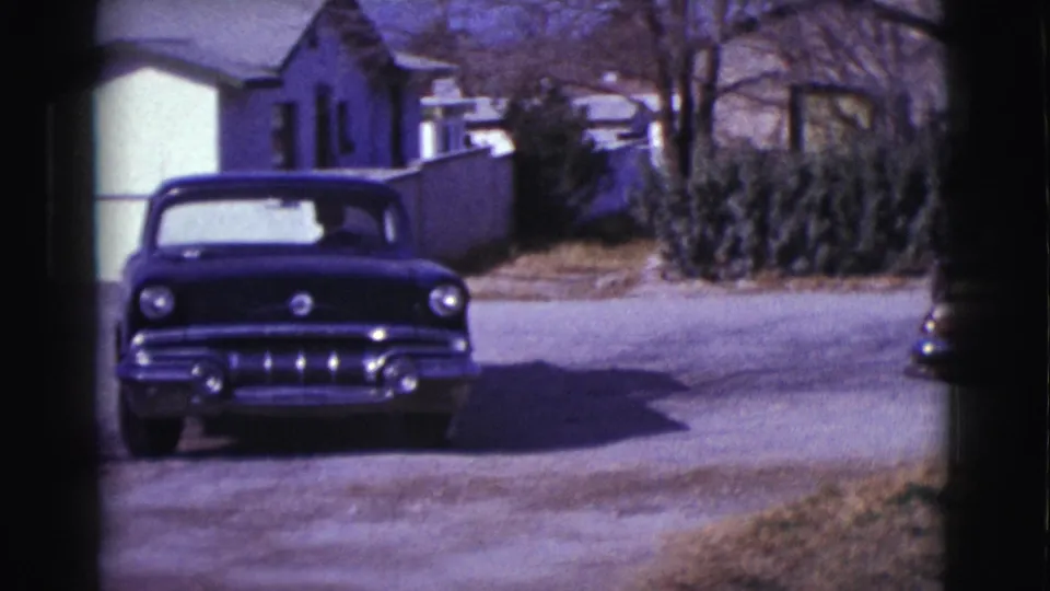 The dark mid-century car is seen from the front at greater distance on a Clarkdale residential street, framed by houses and shrubs.
