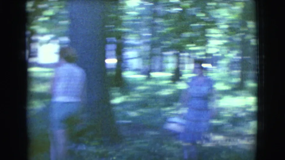 Two women in period clothing walk through a wooded area in 1961 home-movie footage, with one carrying a basket.