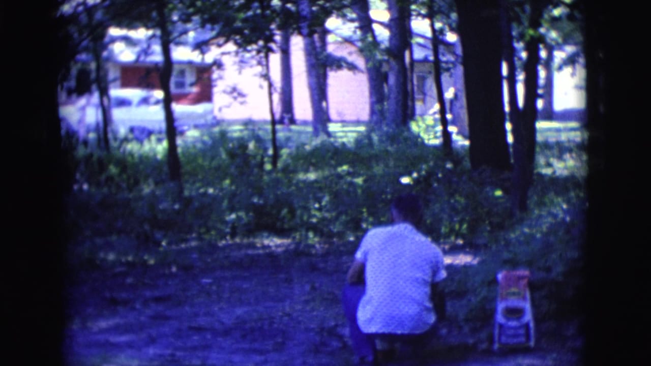 Man crouching in a shaded park near a small child's chair with houses and a car behind trees.