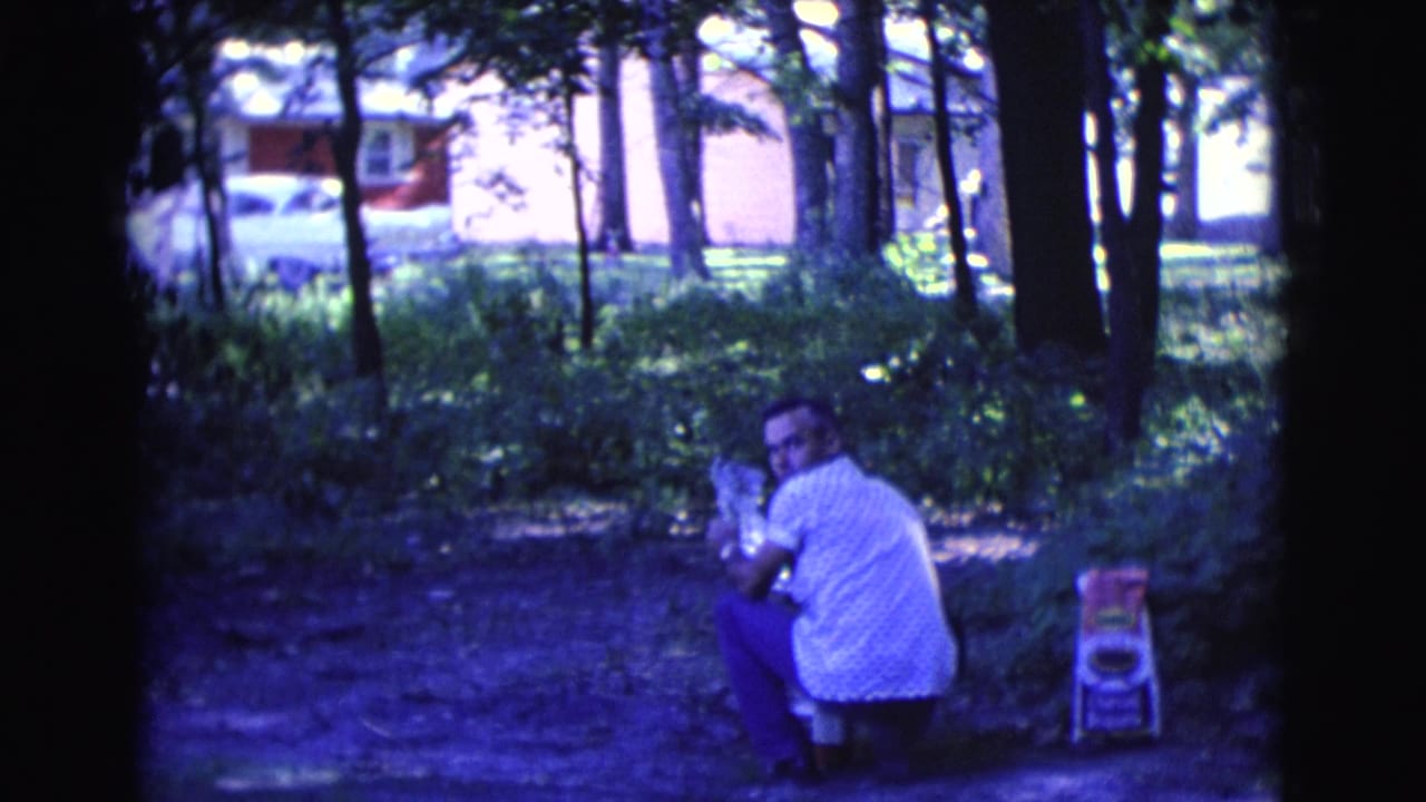 A man in a white shirt turns toward the camera while crouched in a shaded Clarkdale park, with a small chair and residential background visible.
