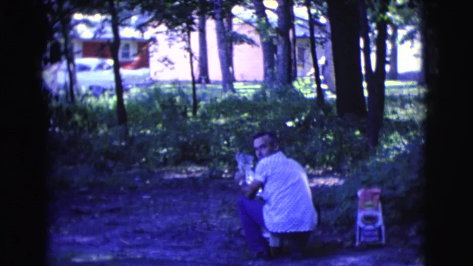 A man in a white shirt turns toward the camera while crouched in a shaded Clarkdale park, with a small chair and residential background visible.