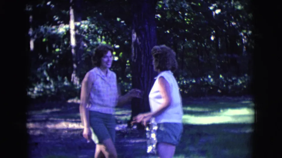 Two women in sleeveless tops and shorts stand in conversation beneath trees in the 1961 Clarkdale home-movie park scene.