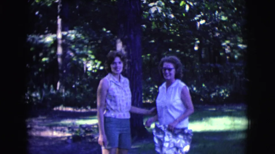 Two women face the camera in a wooded Clarkdale park, wearing light 1960s summer clothing against a shaded green background.