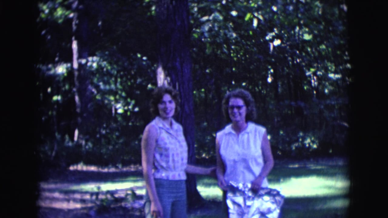 Two young women stand in a wooded grassy area, one wearing glasses and holding a shiny bag.