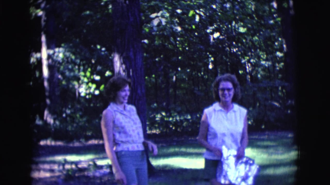 Two women walk and smile in a sun-dappled wooded area.