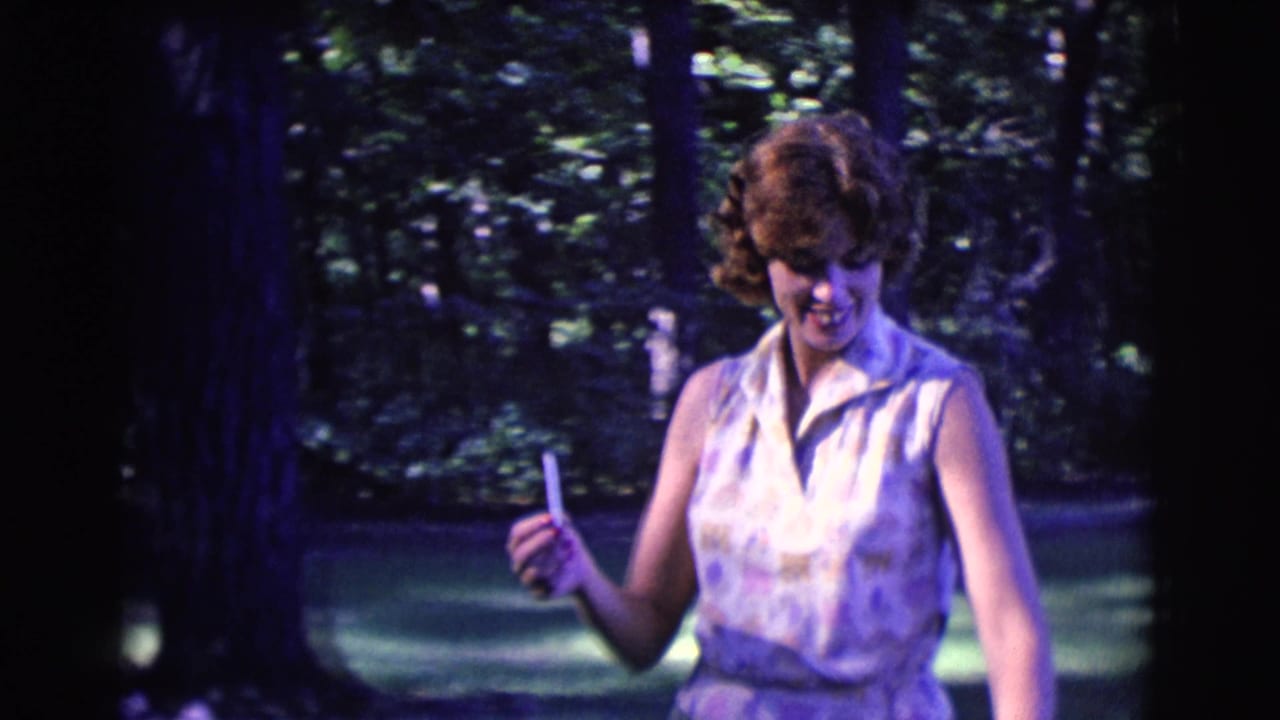 Woman in a sleeveless patterned dress holds a cigarette while standing among trees.