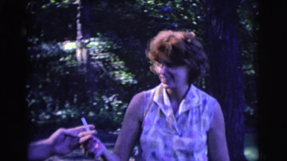 The woman smiles in the wooded setting as another person reaches in to take the cigarette in this 1961 Clarkdale home movie frame.