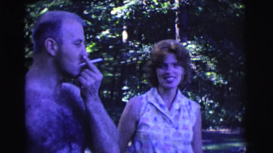 A shirtless man brings a cigarette to his mouth while the woman smiles nearby in 1961 Clarkdale outdoor home-movie footage.