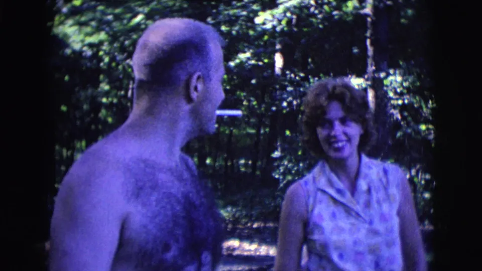 The shirtless man faces the woman with a cigarette between them in a shaded wooded setting from the 1961 Clarkdale home movie.